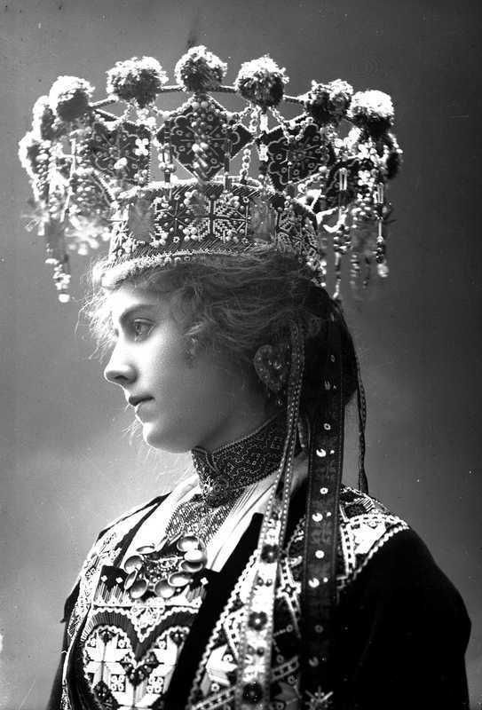 Black and white portrait of a woman in profile wearing elaborate, traditional, ornate folk attire with intricate patterns and a large, decorative headdress adorned with flowers and hanging beads.