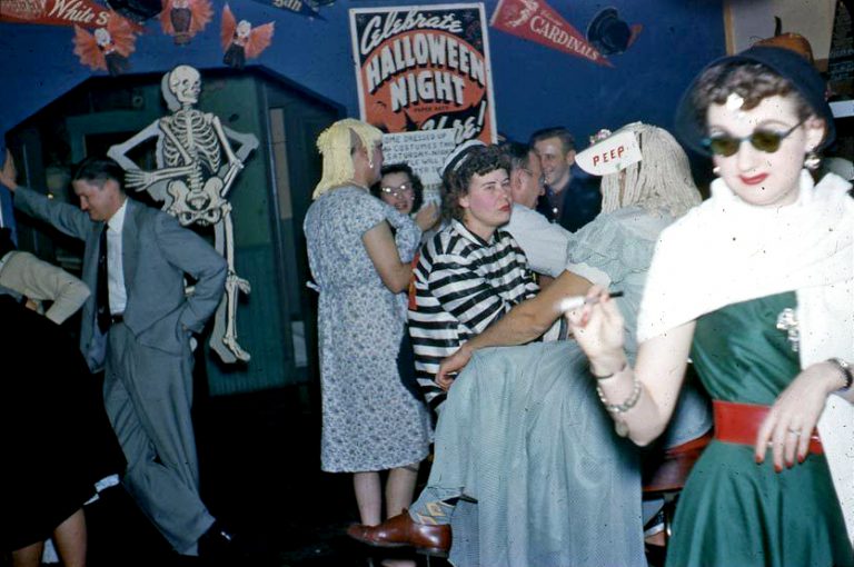 People in vintage costumes gather at a lively Halloween party. Some wear masks, hats, or glasses. A skeleton decoration and a “Celebrate Halloween Night” sign are visible in the festive background.