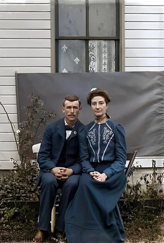 A man and woman in vintage clothing sit side by side on chairs outside a house with a window behind them. They both face forward with neutral expressions, and dried plants are visible near their feet.