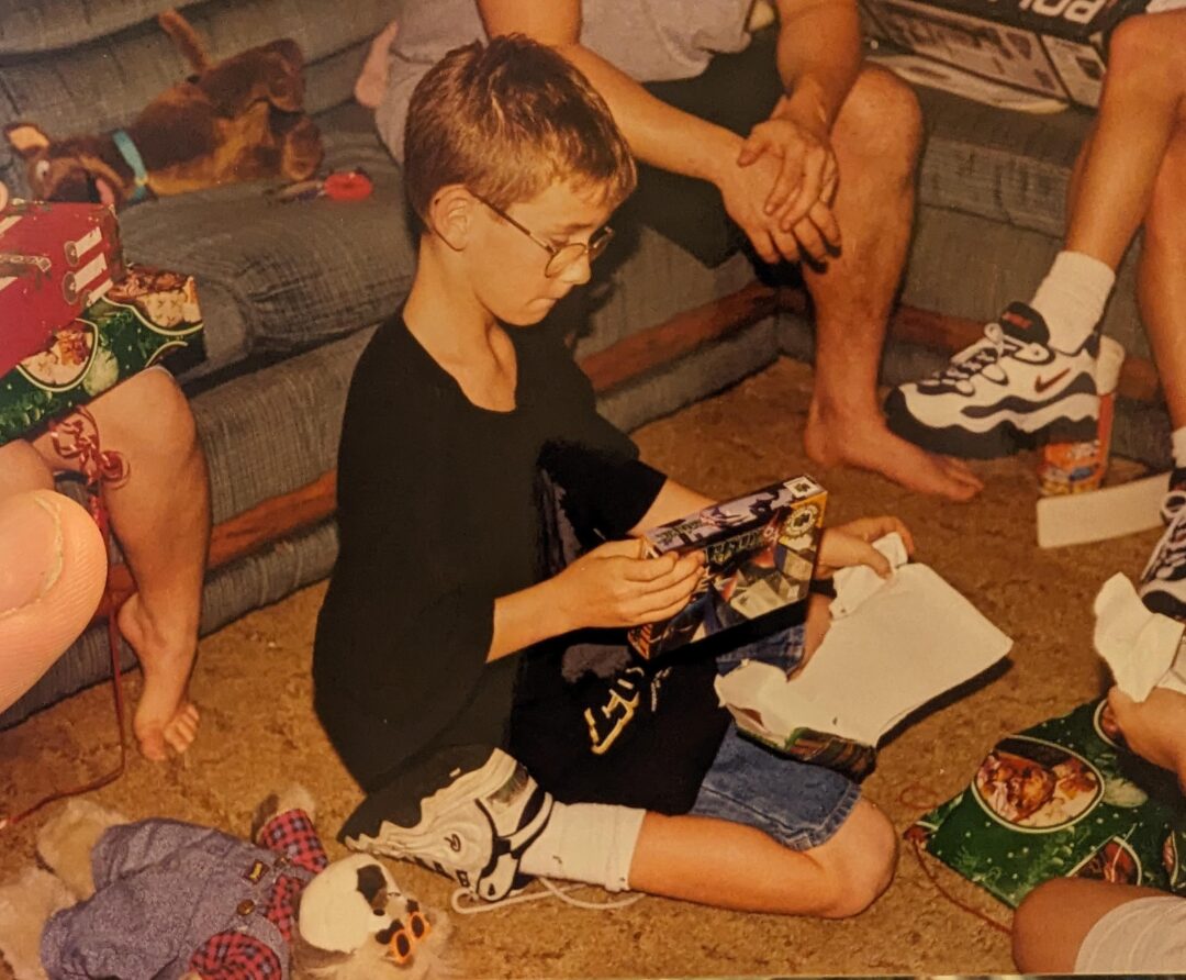 A boy wearing glasses sits on the floor opening a gift, surrounded by people and snacks. He appears focused on the present, while others, mostly off-frame, watch or sit nearby on a couch. Holiday toys and snacks are scattered around.