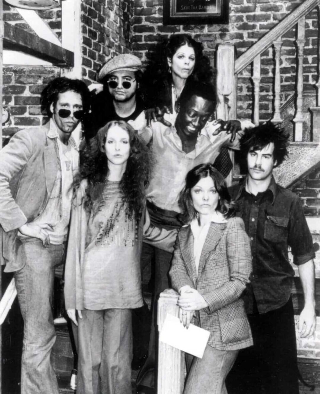 A group of seven people poses together on a staircase in front of a brick wall. Some are standing, others sitting or leaning on the railing, all wearing 1970s-style clothing and serious or playful expressions.
