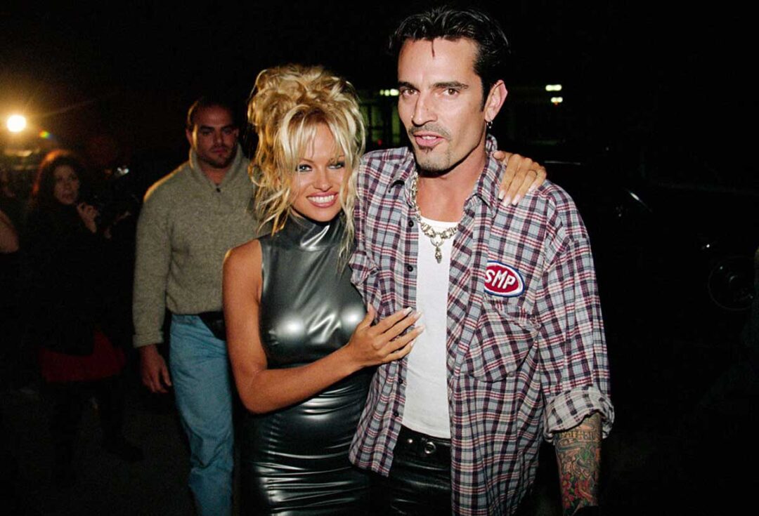 A blonde woman in a shiny silver dress hugs a man wearing a plaid shirt, white T-shirt, and necklaces. They are walking outdoors at night with other people and lights in the background.