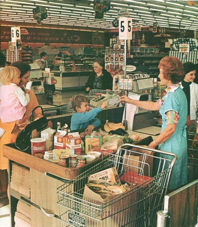 A woman with two children checks out groceries at a supermarket counter, handing money to a cashier. Several food items fill the cart, and other customers and employees are visible in the background.
