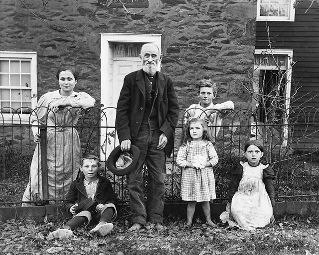 A black-and-white photo of an older man holding a hat, standing in front of a stone house with two women and three children, all posing by a metal fence. The adults stand behind the fence while the children sit or stand in front.