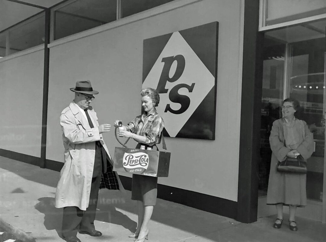 A woman hands a man a Pepsi-Cola bottle outside a store with a large "ps" sign, while another woman stands smiling by the entrance. The scene appears to be from the mid-20th century.