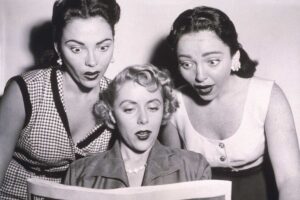 Three women with surprised expressions look at a newspaper together; two stand on either side of the woman sitting and holding the paper. The image appears to be from the mid-20th century.