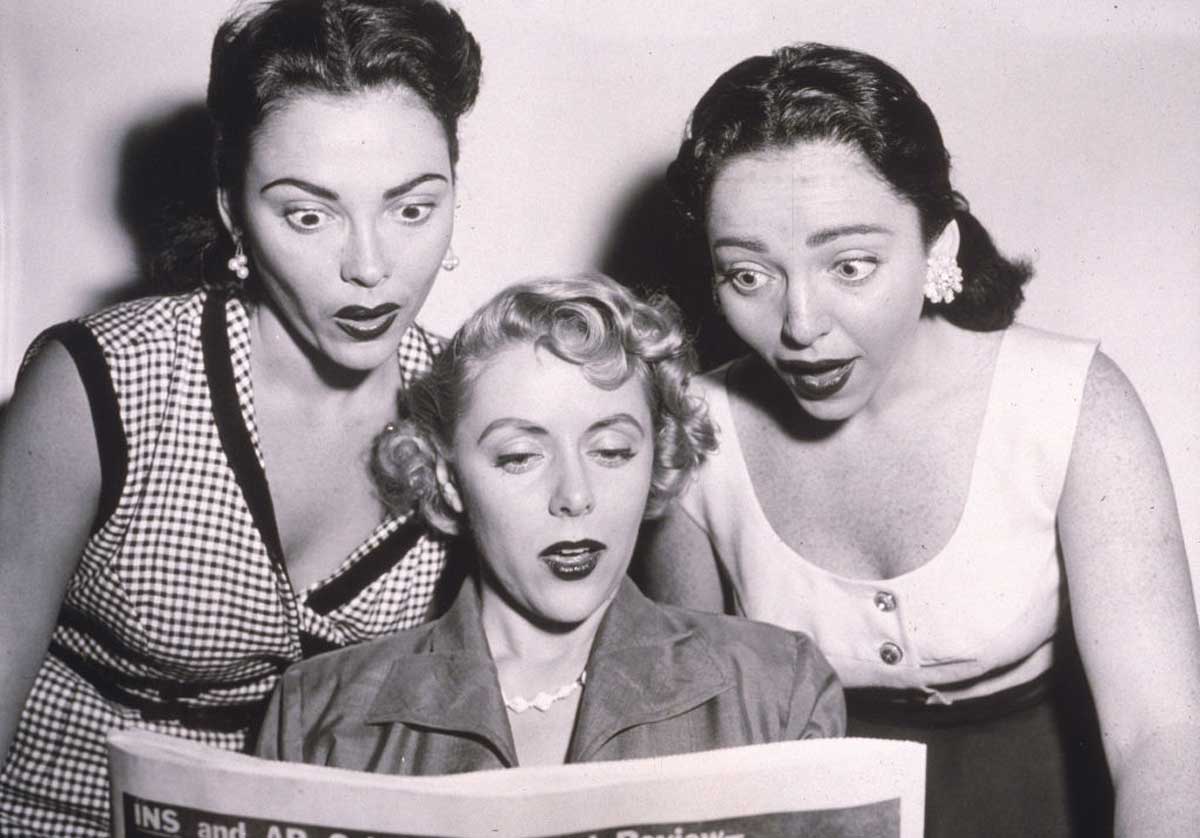 Three women with surprised expressions look at a newspaper together; two stand on either side of the woman sitting and holding the paper. The image appears to be from the mid-20th century.