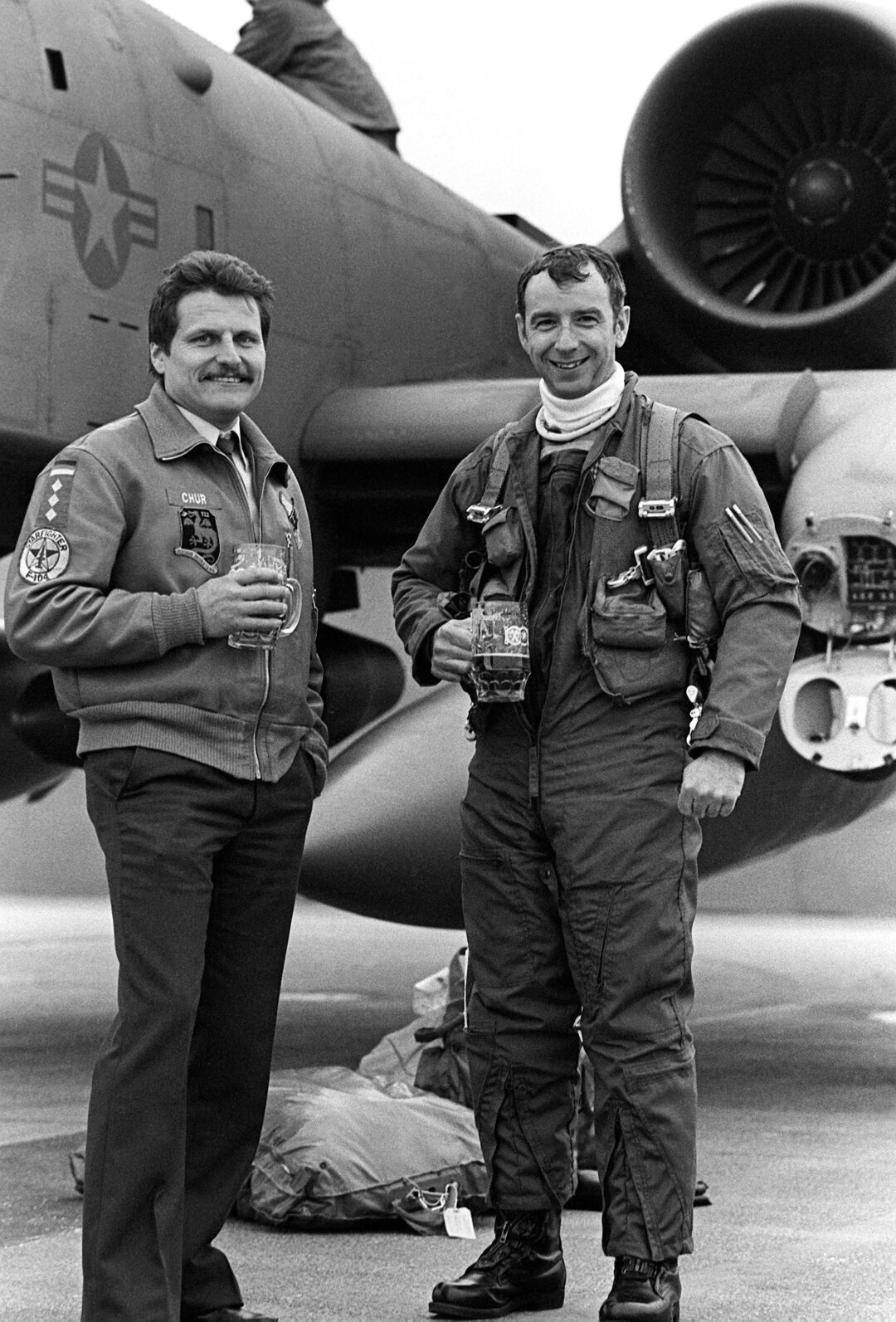 Two men in flight suits stand smiling in front of a military aircraft, holding drinks. One wears a jacket, the other a flight suit. A jet engine and aircraft tail are visible in the background.