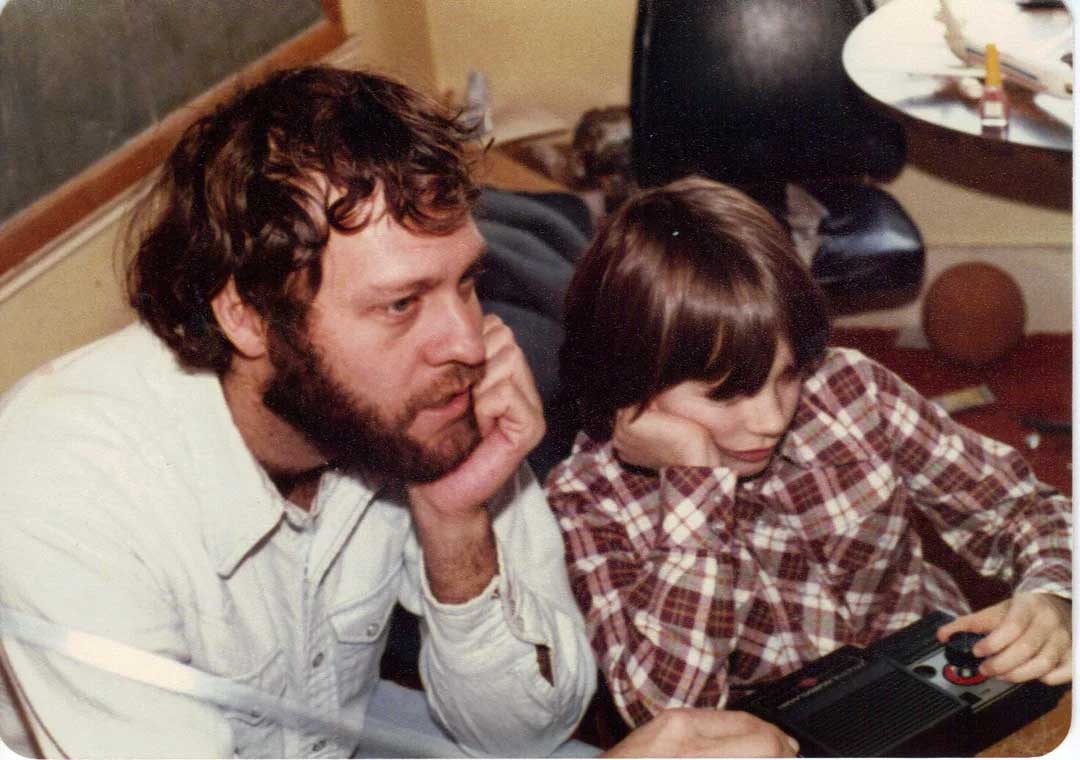 An adult man and a child sit closely together, both resting their chins on their hands, looking intently at a video game console controller that the child is holding. They appear focused and engaged.