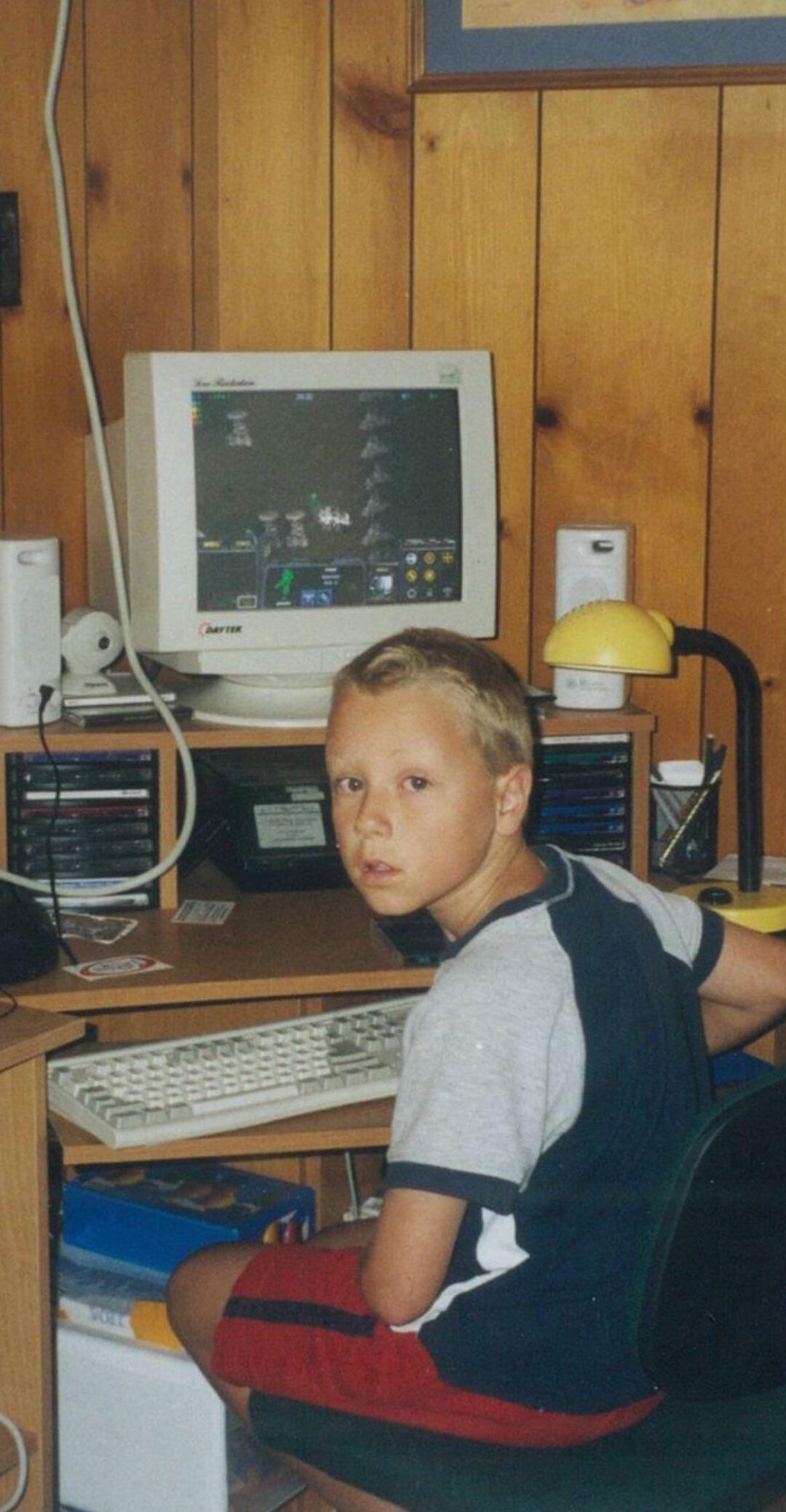 A young boy sits at a wooden desk using a large, old computer monitor. He looks toward the camera, appearing surprised. The desk has CDs, speakers, a lamp, and wood-paneled walls in the background.