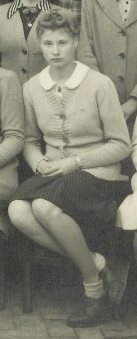A young woman with short, wavy hair sits on a chair, legs crossed. She wears a light cardigan with a ruffled front, a collared blouse, a striped skirt, socks, and dark shoes. The image is black and white.