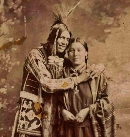 A vintage sepia photograph of a Native American man in traditional regalia with feathers and a woman in patterned clothing, standing close together and smiling at the camera. The man has his arm around the woman's shoulders.