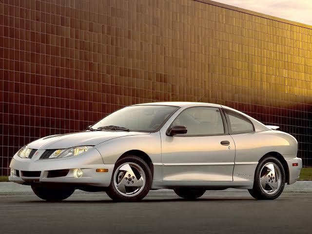 A silver two-door coupe car is parked on a paved surface in front of a modern brown tiled building during daylight.