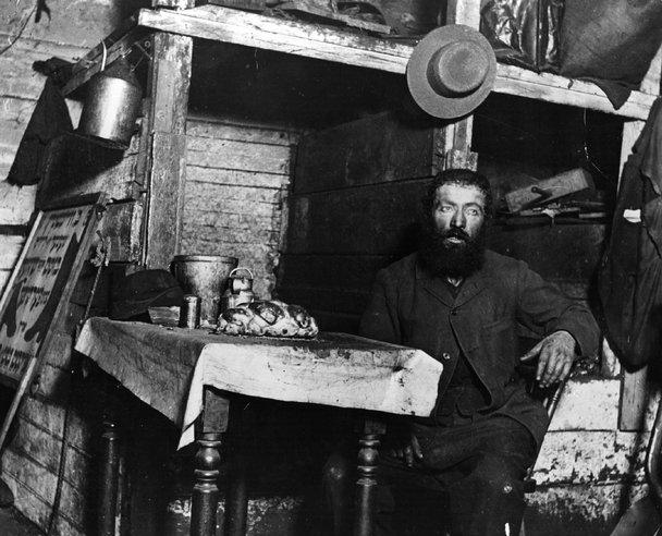 A bearded man sits at a small table covered with a cloth and food in a rustic, cluttered wooden room. A hat hangs on the wall above him, and shelves and household items surround the scene.