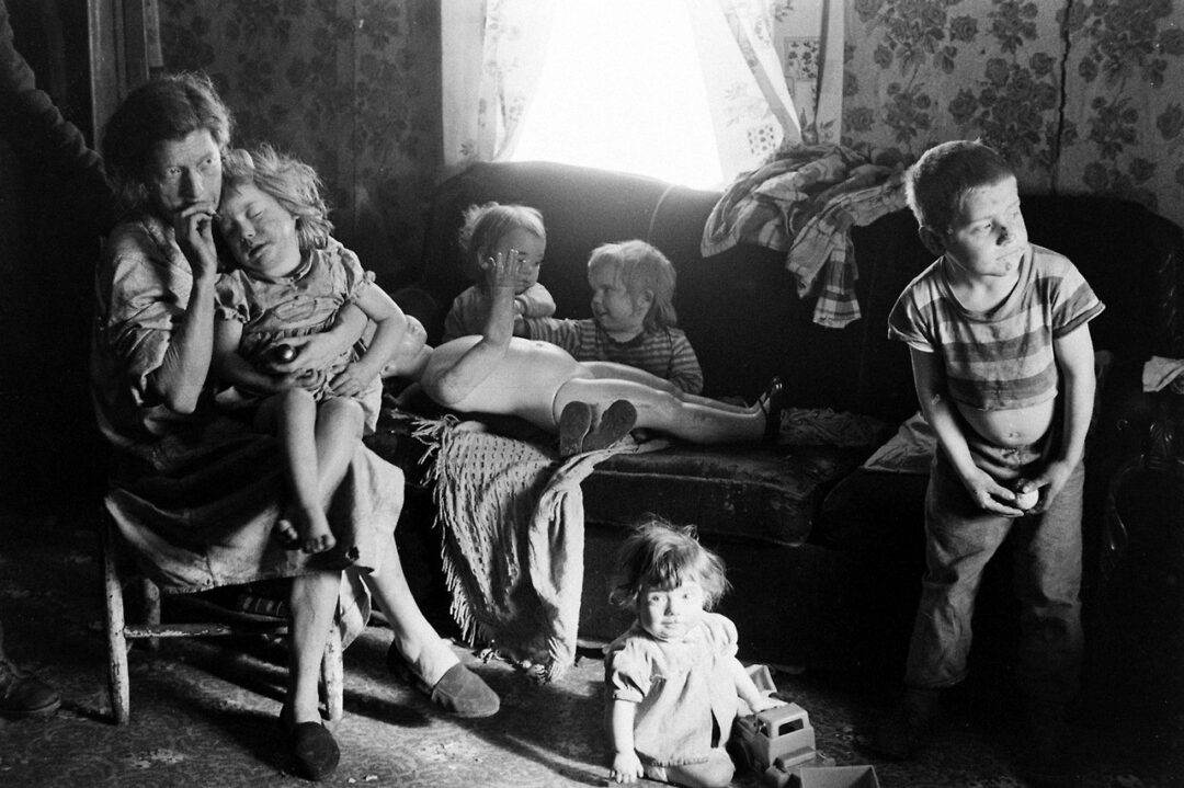 A woman sits on a chair holding a young child, with three more children on a couch and one toddler on the floor playing with a toy; sunlight streams through a window behind them in a modest, cluttered room.