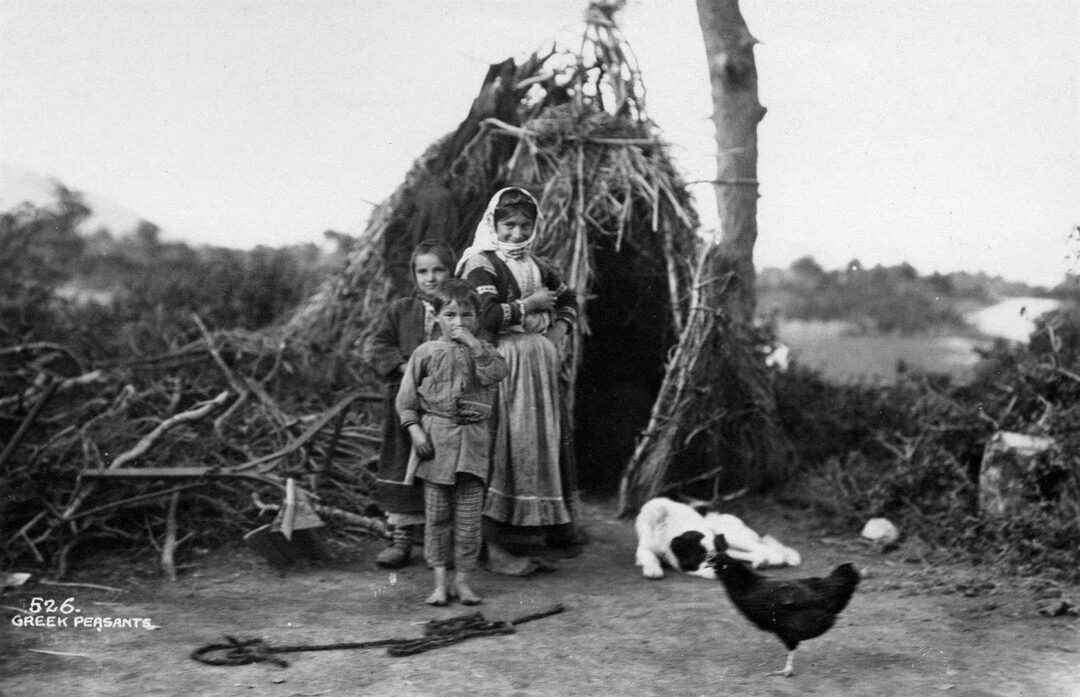 Three people, including two children and an adult, stand in front of a rustic hut made of branches. A dog lies on the ground and a chicken stands nearby. The background shows trees and open countryside.
