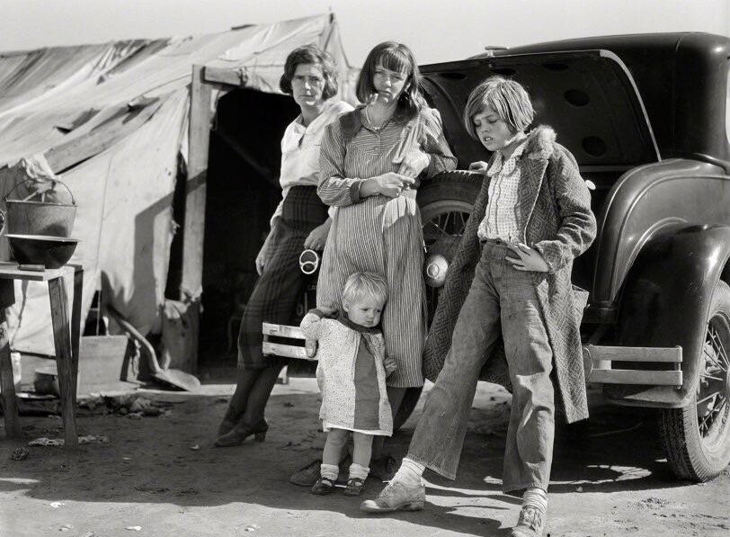Four children stand in front of a car with an open trunk, next to a canvas tent. Their clothing is worn and the setting appears dusty, suggesting hardship during the Great Depression era.