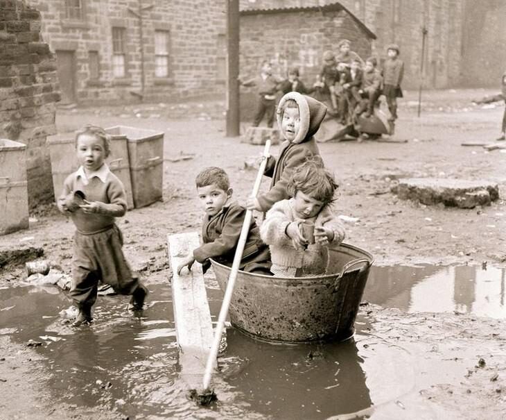 Four children play in a muddy, puddle-filled courtyard; two sit in a large tub using sticks to paddle, while two others stand nearby. Old stone buildings and more children are visible in the background.