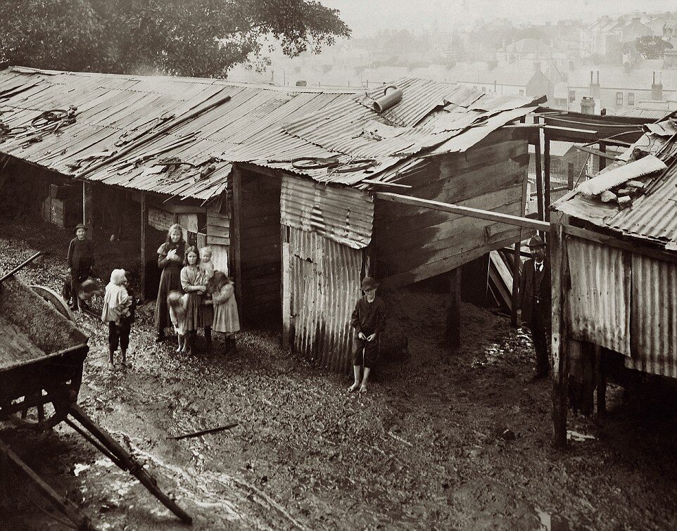 A group of children stand in muddy ground outside makeshift shacks with corrugated metal roofs and walls, in a poor, urban area. The background shows a foggy cityscape and more simple structures.
