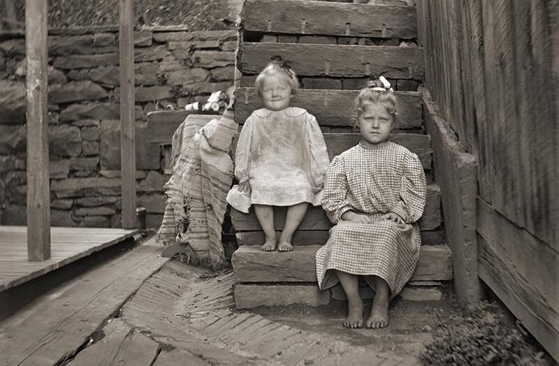 Two young girls in old-fashioned dresses sit barefoot on stone steps outside, next to a wooden fence and a brick wall. One girl smiles, while the other looks serious. A striped blanket rests on the steps beside them.