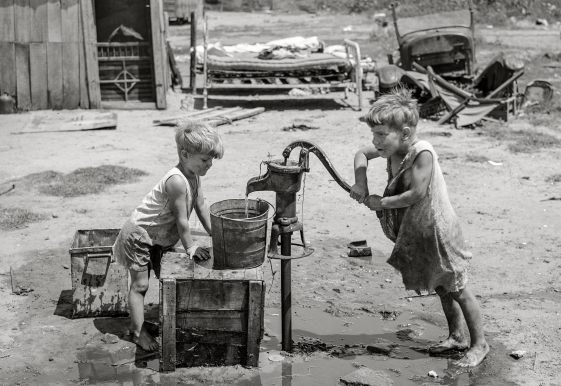 Two young children in worn clothing use a hand water pump outdoors, surrounded by mud, crates, and scattered debris. In the background are a broken bed frame and dilapidated wooden structures.