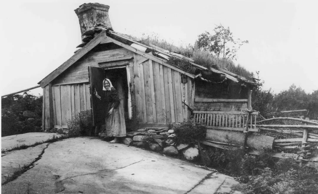 An elderly woman stands in the doorway of a rustic wooden house with a grass-covered roof, surrounded by rocks and simple wooden fencing in a rural, natural setting.