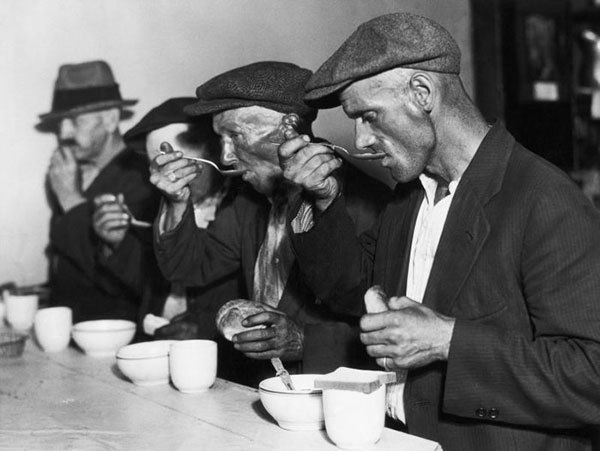 Four men in worn clothing and hats eat soup and bread at a counter, appearing focused on their meal. The black and white photo suggests a historical setting, likely during a time of hardship or economic struggle.