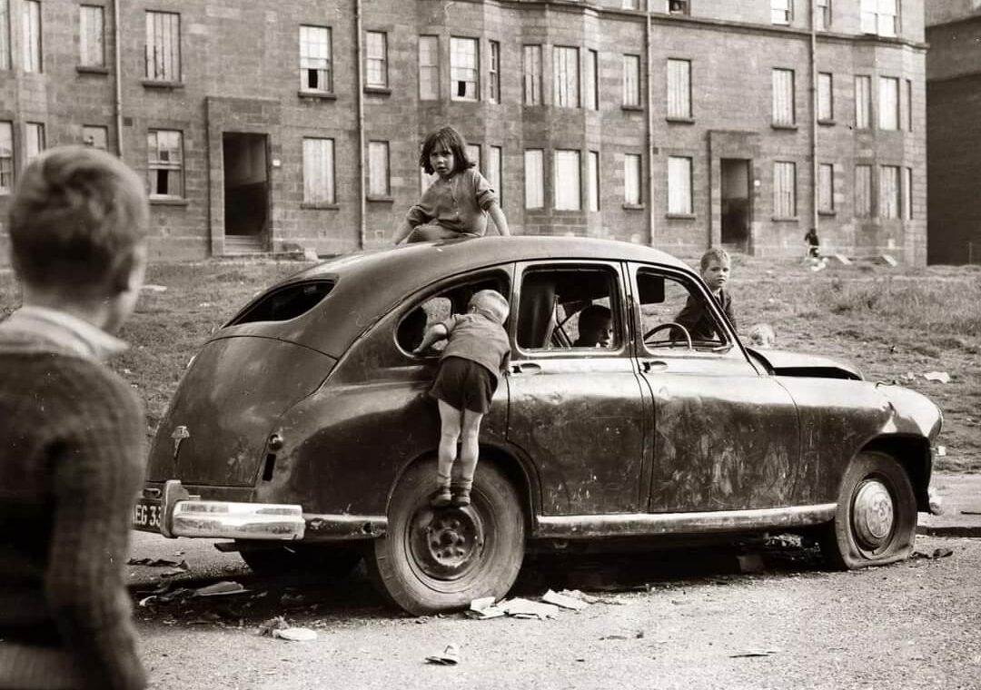 Children play on and inside an old, beaten-up car in a run-down urban courtyard, with apartment buildings in the background. One child climbs through a car window, while others watch and join in.