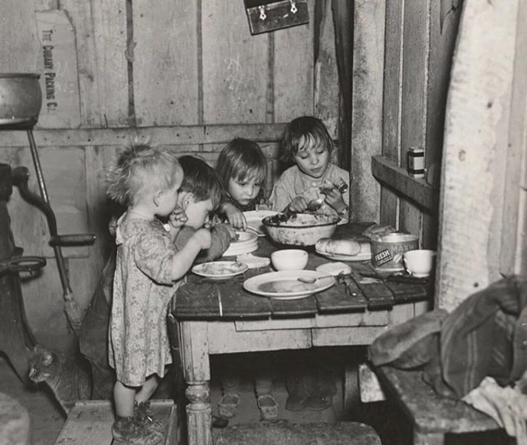 Four young children sit around a small, worn table in a rustic, wooden room, eating a simple meal. The scene looks old-fashioned and modest, with basic dishware and an overall atmosphere of poverty.