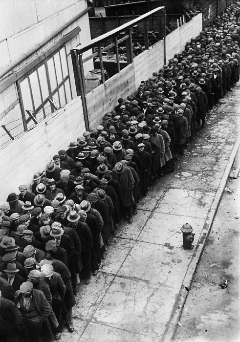 Black-and-white photo of a long line of men in coats and hats standing on a city sidewalk, waiting next to a wooden fence, likely during the Great Depression.