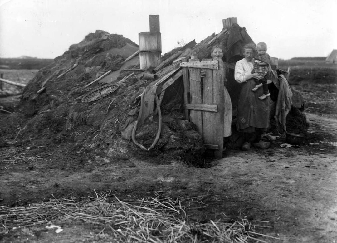 Three people, including a child, stand outside a small, rough shelter made of earth and wood in a rural setting, with scattered debris and barren land around them.