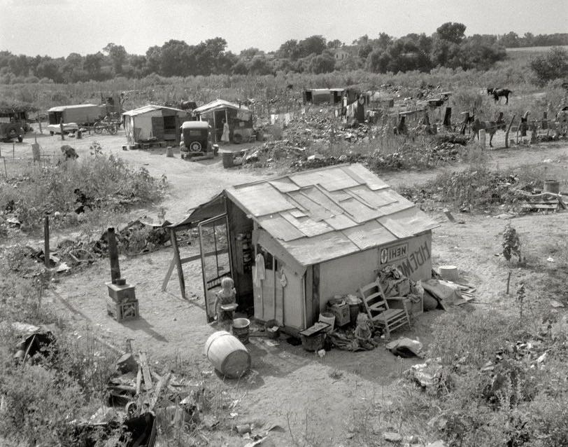 A black-and-white photo shows a small, makeshift shack surrounded by scattered debris and items. Other shacks, vehicles, and people appear in the background, set in a rural, overgrown area.