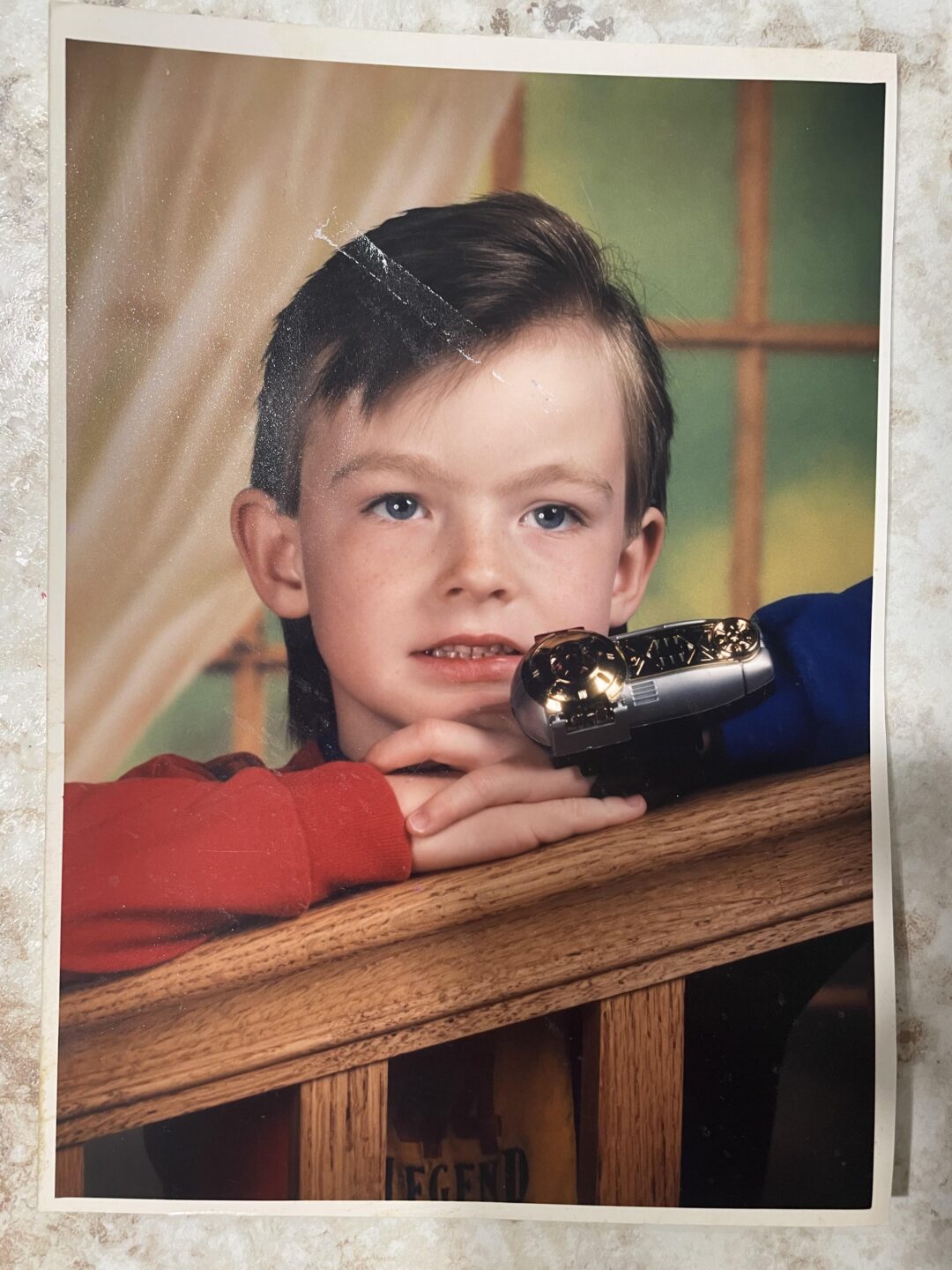 A young boy with light skin and brown hair rests his arms on a wooden railing, looking slightly off camera. He wears a red and blue shirt and holds a toy car. The background has soft, neutral colors.