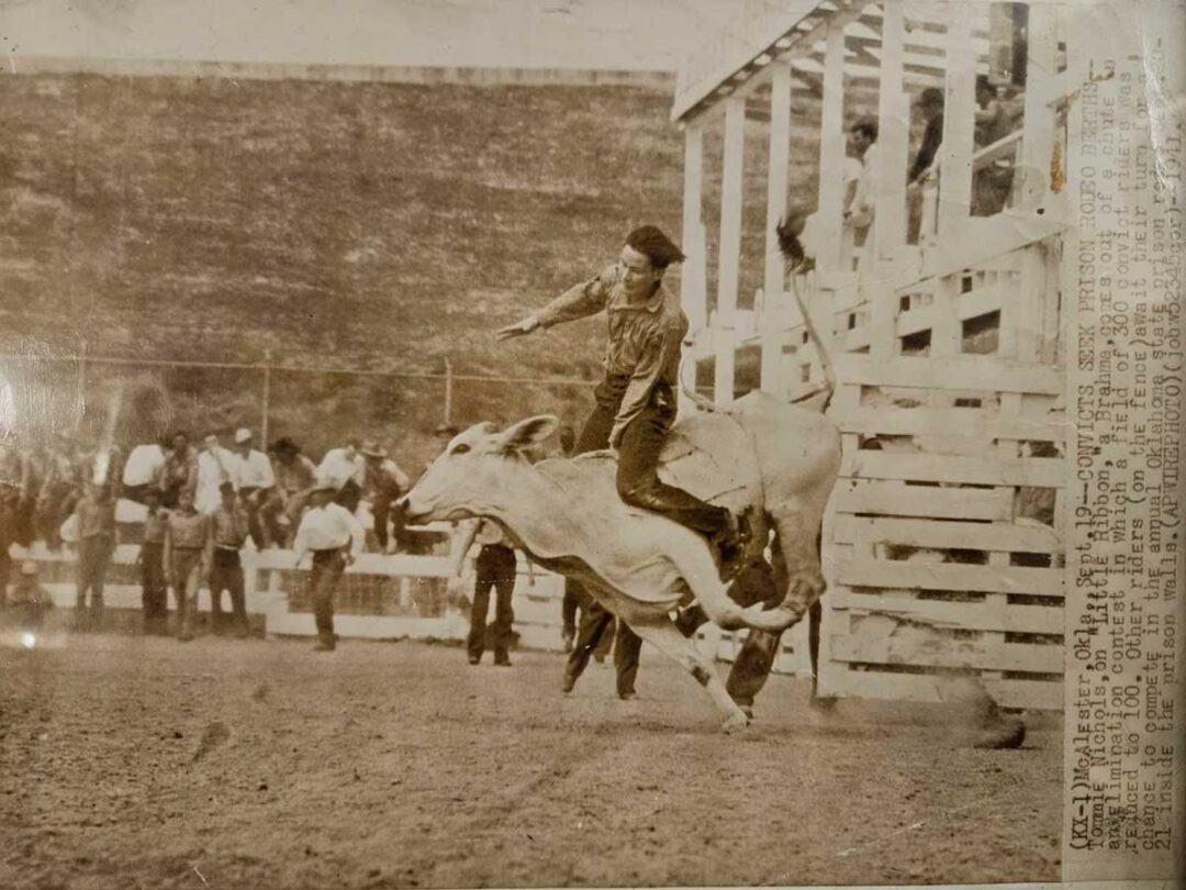 A rodeo rider in mid-action rides a bucking bull in a dirt arena. Spectators watch from behind a white wooden fence, and a few people stand on a raised platform nearby. The photo has a sepia tone, indicating it is historical.