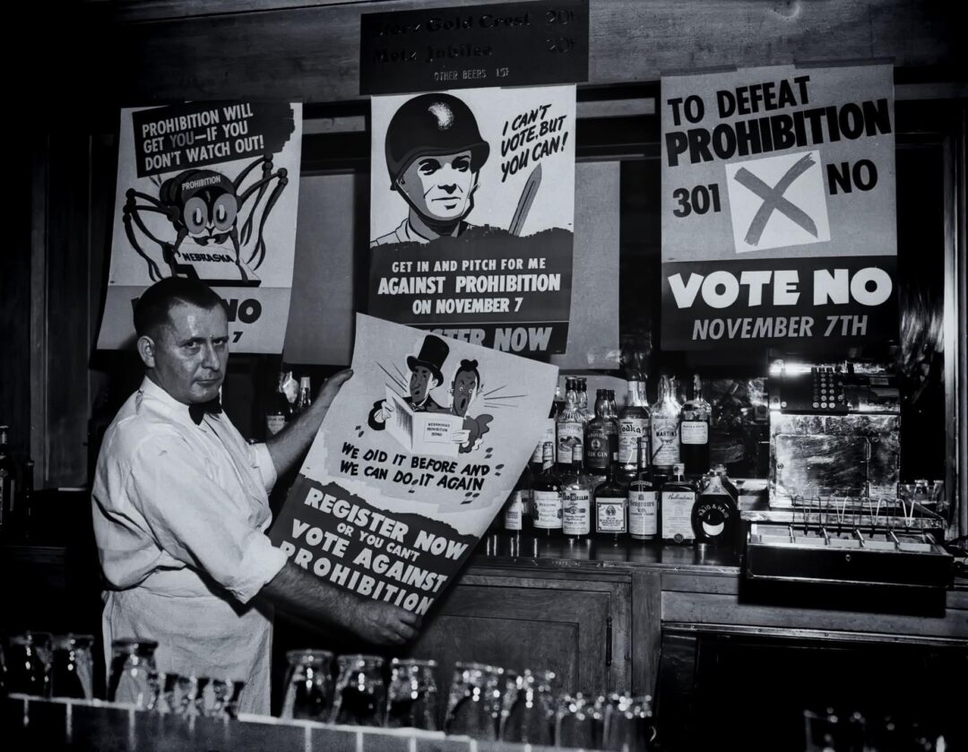 A bartender stands behind a bar holding an anti-prohibition poster, surrounded by liquor bottles and more signs urging people to vote against prohibition on November 7.