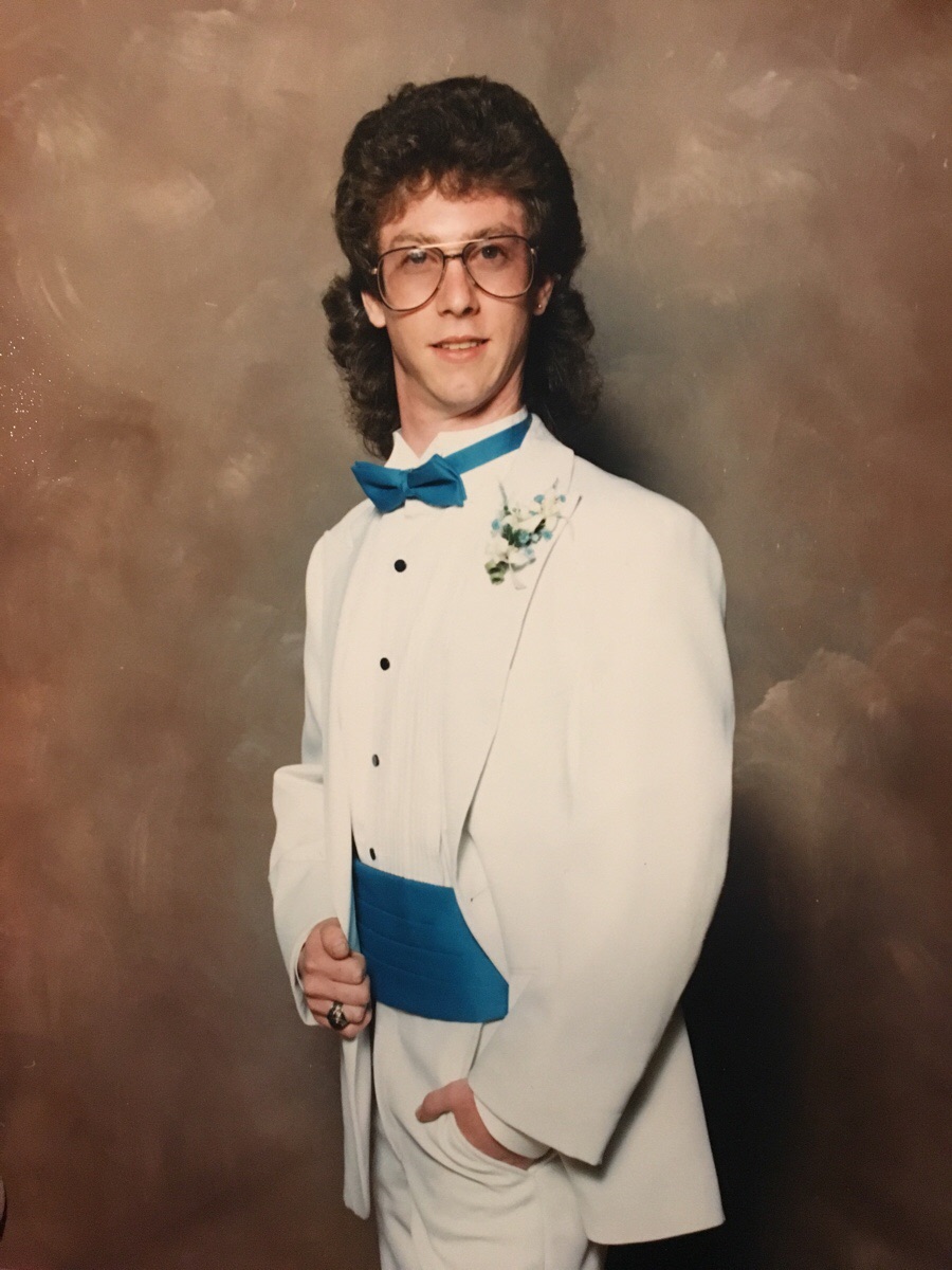 A young man wearing a white tuxedo with a blue bow tie and cummerbund poses for a formal portrait against a brown studio backdrop. He has curly hair, glasses, and is holding his jacket with one hand.