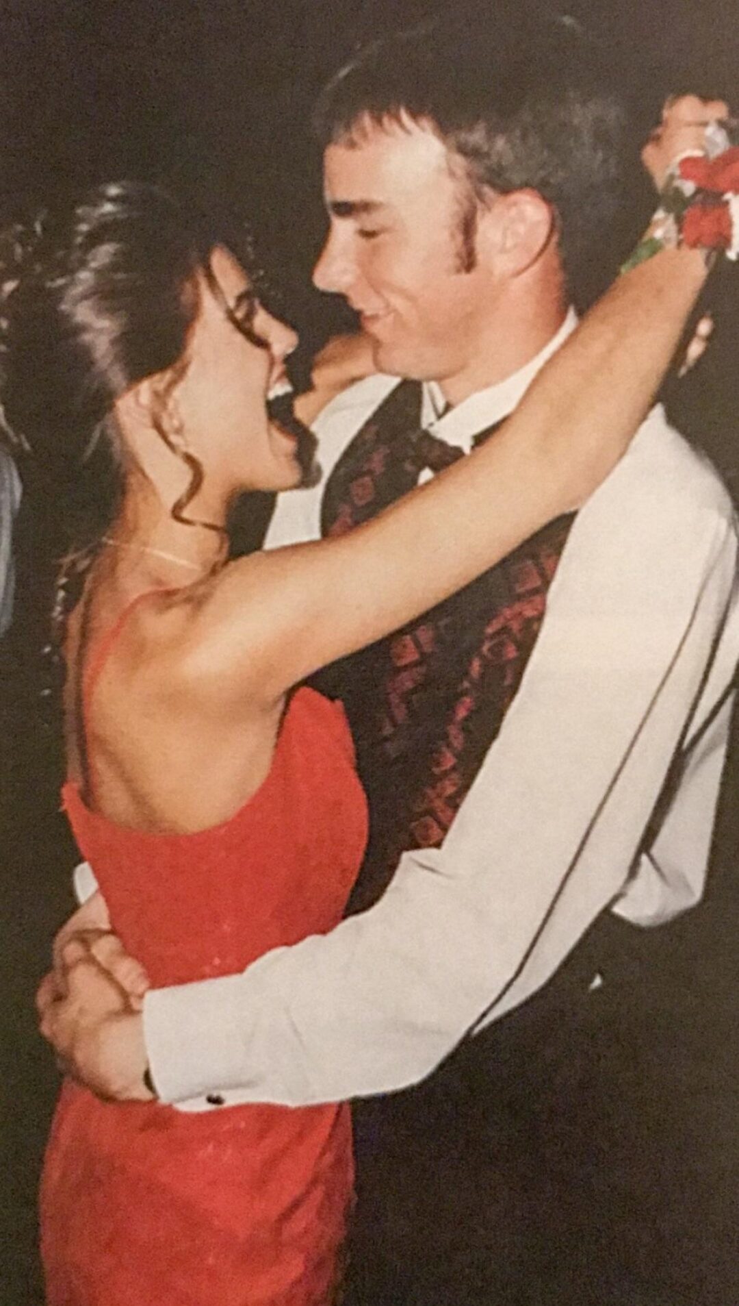 A young couple dressed formally, smiling and holding each other while dancing closely. The woman wears a red dress, and the man is in a white shirt with a patterned vest and tie.