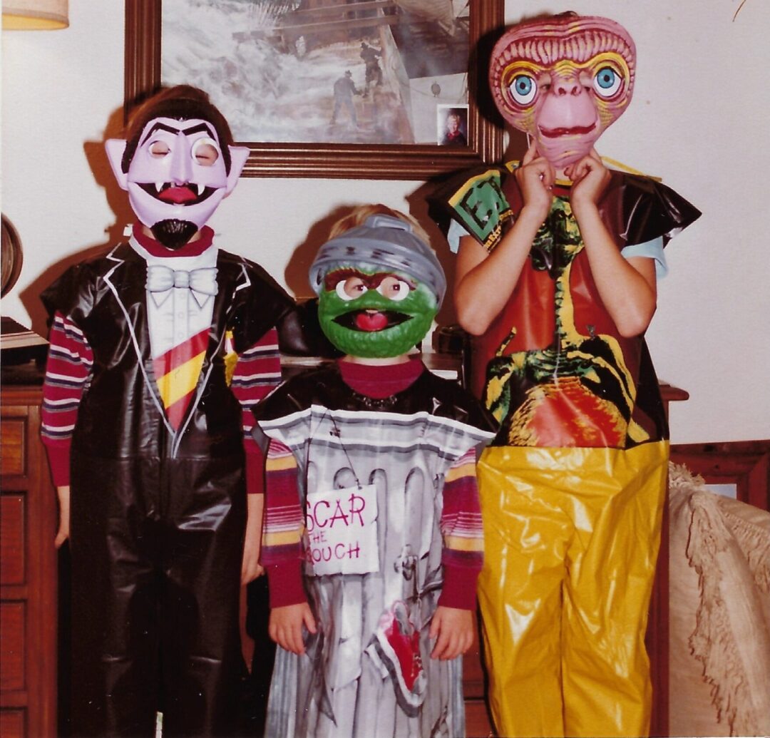 Three children in colorful Halloween costumes and masks stand indoors in front of a painting of a sailing ship. One is dressed as Count von Count, another as Oscar the Grouch, and the third as E.T.