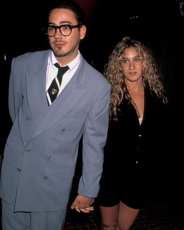 A man in a light blue double-breasted suit and glasses holds hands with a woman in a black blazer dress with curly blond hair; both stand indoors and look in different directions.