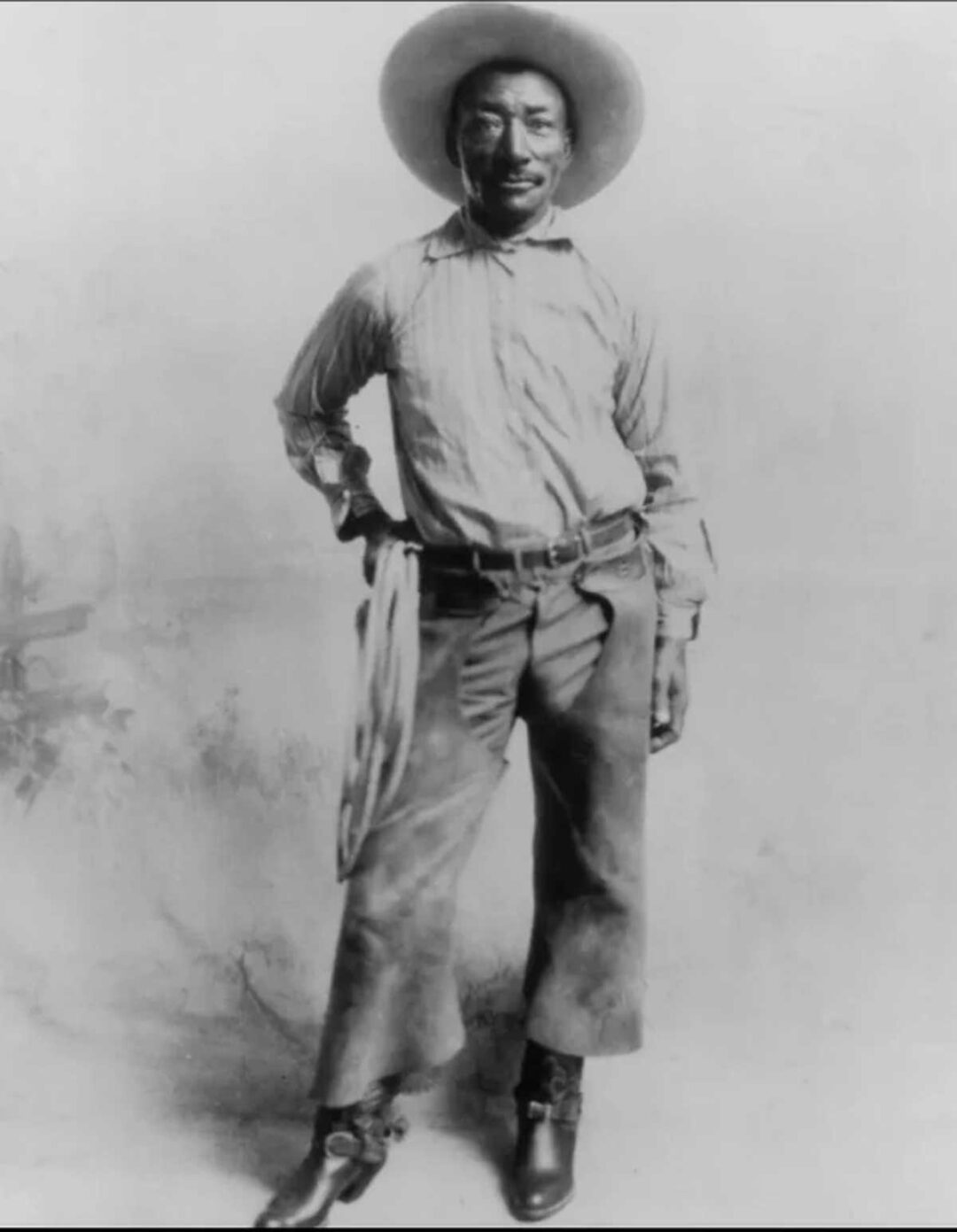 Black-and-white photo of a Black cowboy standing confidently, wearing a wide-brimmed hat, striped shirt, chaps, boots, and a scarf tied at his waist, with one hand resting on his hip.