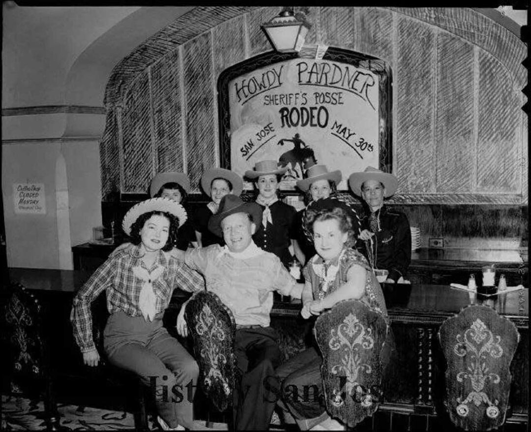 A group of people dressed in cowboy and cowgirl outfits sit and stand at a bar, smiling. Behind them is a large sign reading "Howdy Pardner Sheriff's Posse Rodeo San Jose May 30th.
