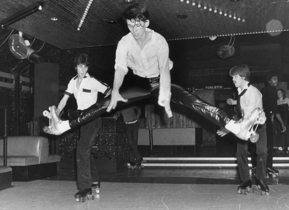 A man on roller skates leaps into a high split jump, while two other skaters watch him in a roller disco setting with a raised stage and disco lights.