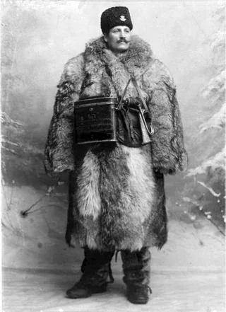 A man stands wearing a thick fur coat, boots, and a fur hat with a badge. He holds a metal canteen and a small satchel, posing in front of a snowy, winter-themed backdrop in a black and white photo.