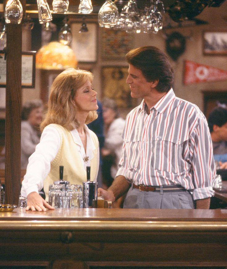 A woman and man stand at a bar, facing each other and smiling. The woman wears a cream vest over a blouse; the man wears a striped shirt. Glasses hang overhead, and the background shows a busy, warmly lit bar scene.