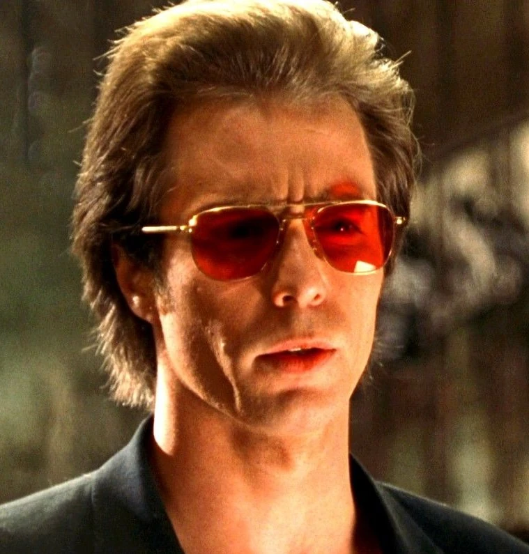 A man with light brown hair wearing red-tinted sunglasses and a black jacket looks serious, standing outdoors with a blurred background.