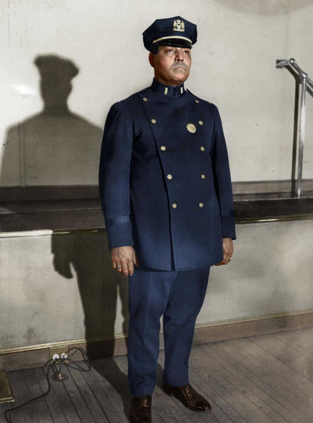 A Black police officer in a formal blue uniform and cap stands indoors in front of a light-colored wall, casting a shadow behind him. The floor is wooden and there is a metal railing to the right.