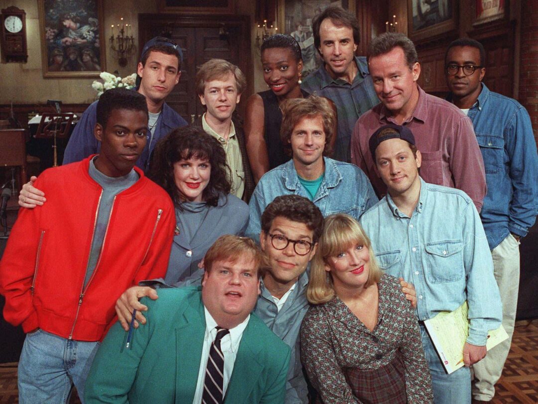 A group of thirteen people, including men and women, pose closely together indoors, smiling at the camera. The group is diverse in age, race, and style, with casual clothing and a lively, friendly atmosphere.