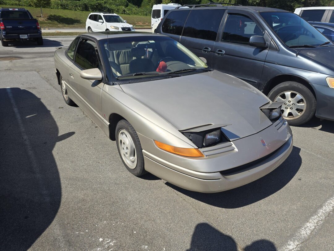 A beige Saturn coupe with one pop-up headlight stuck open and the other partially closed is parked in a parking lot between two other vehicles on a sunny day.