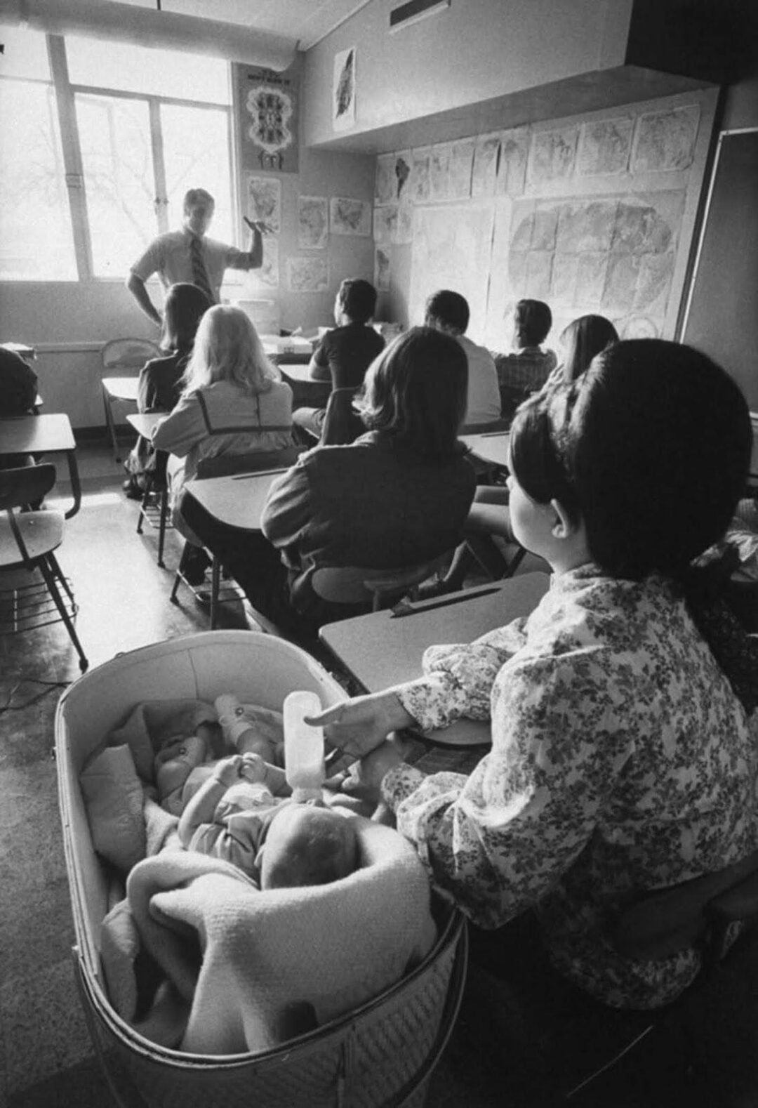 A woman sits in a classroom feeding a baby from a bottle in a bassinet while listening to a teacher who stands at the front; other students sit at desks facing the teacher.