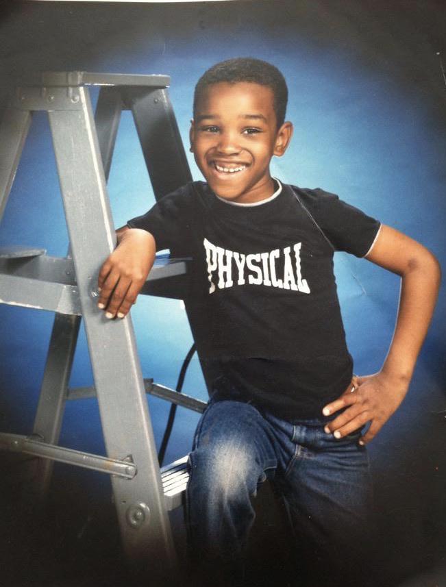 A smiling young boy wearing a black t-shirt with the word "PHYSICAL" and blue jeans poses with one arm resting on a metal ladder against a blue studio backdrop.